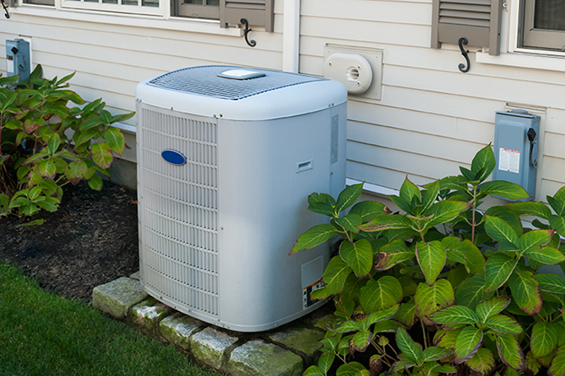 An outdoor air conditioning condenser unit installed on a paver base next to the exterior wall of a home with light-colored siding and surrounding green foliage.